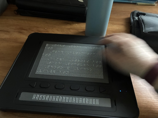 This is a picture of the dot pad. It is a black plastic device and at the top there is a large square area that is 10 lines long by 30 characters wide. Below that there is a divider with two arrow shaped buttons that are for panning, the display and four oval shaped function keys. Below that there is the traditional single line of braille. The single line of braille is used for looking at captions and the larger area can be used for looking at pictures, or you can just use the larger area to read multiple lines at once rather than having to continuously scroll