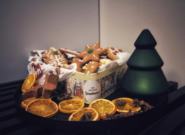 A box of decorated "pepperkaker" (norwegian gingerbread cookies) on a black round plate on a table, decorated with dried orange slices and a little Christmas tree made of frosted glass