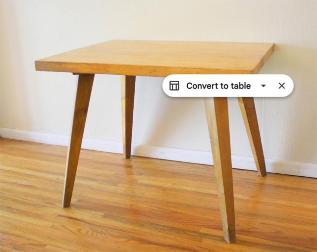 A wooden table has a Google Sheets LLM prompt saying “Convert to table” overlaid on its right hand side. The table stands on a natural wood floor and next to a white wall.