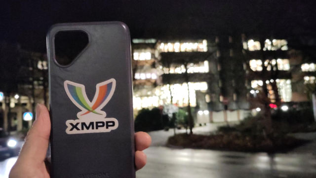 A picture showing a bumper case of a fairphone with an XMPP sticker attached to it. In the background there is the library of the University of Oldenburg. The picture was taken at night 