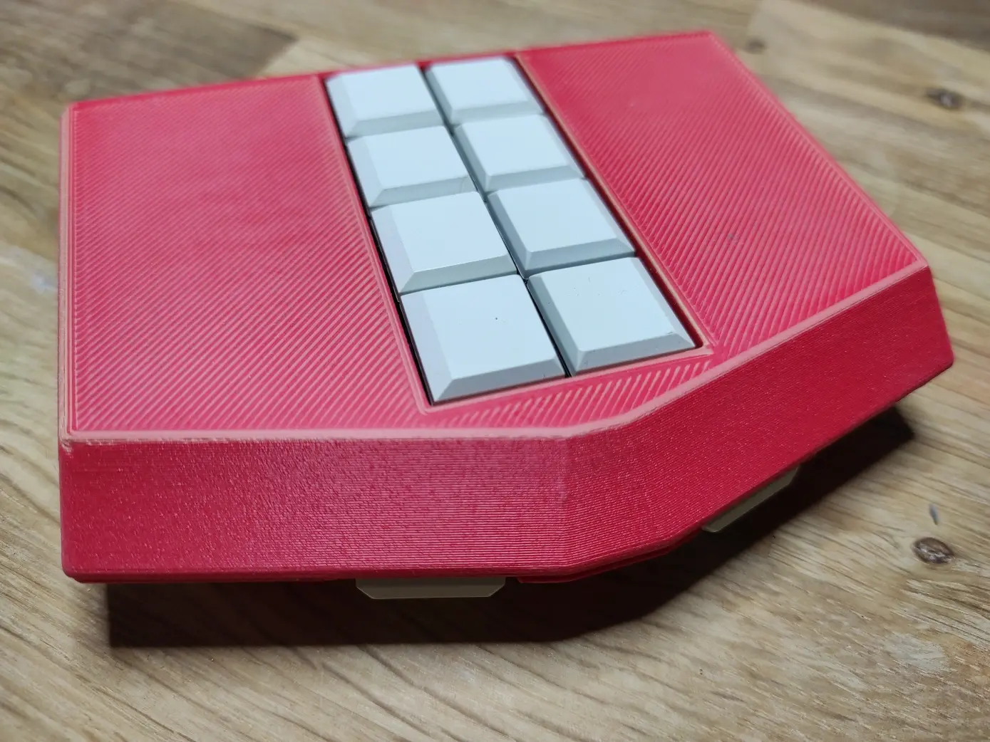 Mobile Braille-Tastatur Metabraille mit 8 Tasten auf der Vorderseite und zwei seitlich sichtbare Tasten auf der Oberseite. Das Gehäuse ist 3D-gedruckt, rot mit hellen Streifen und die Tasten sind weiß. Das Gehäuse ist ungefähr fünfeckig. Ein Rechteck wird an der Breitseite durch ein Trapez erweitert. Die Form ähnelt einem Haus mit Fenstern in der Mitte. Die Tastatur zeigt mit der Oberseite nach rechts unten und liegt auf einer Holzplatte.