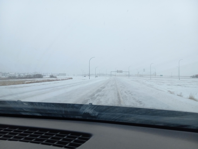  a view through the passenger side of the windshield of a car on a road that is completely coated in snow.  If you look carefully, you can sort of see where tire tracks are.  Otherwise the road is pretty indistinguishable from the rather flat land on either side of the road.  It's snowing and the sky is cloudy.