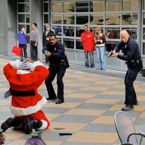 Two policemen with drawn sidearms face a person in a Santa costume on their knees, hands on their head. 