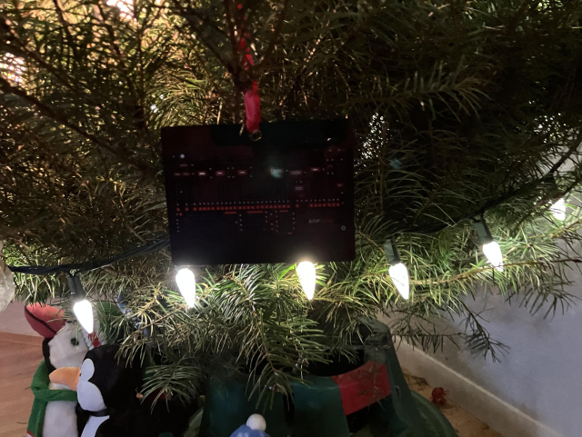 Image shows Christmas tree branches with lights on. There is a flat metal rectangular black ornament that appears to have knobs and sliders drawn on it. Text reads Arp 2600.