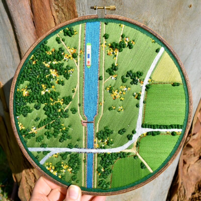 A hand holds a circular embroidery hoop showing a detailed, textured aerial landscape with fields, roads, clusters of green trees, and a bright blue canal running vertically through the center. A small embroidered boat floats on the canal, adding a pop of color against the green terrain. The stitching varies in texture and density to mimic foliage, paths, and water, giving the scene a rich, tactile depth