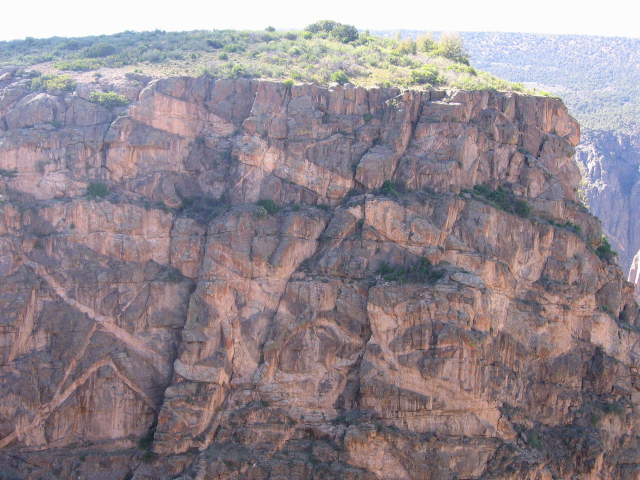 A wall of rock exposed in the Black Canyon of the Gunnison in western Colorado. The wall consists of dark grey-ish, Precambrian-age gneiss and schist dissected by bands of pink, granitic, also-Precambrian-age pegmatites (igneous intrusions that were injected into fractures in the gneiss long after it formed and while it was all still buried deep underground). The metamorphic rocks formed around 1.8 billion years ago, but existed as sedimentary rocks of undetermined age before being metamorphosed. The pegmatites, which include crystals upward of 2 m (6 ft.) in length, are a bit younger at around 1.35 billion years old. The green plant life at the top of the wall grows on mudstone exposures of the Upper Jurassic (~150 million year old) Morrison Formation, which has produced a few dinosaur fossils in the park.