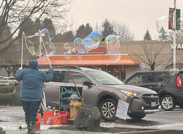 Person standing on a corner in a busy retail development, holding up two sticks with rope between them, that have been dipped in bubble soap and create these massive floaty, rolly bubbles set free into the roadway, shiny iridescent blobs of joy.
