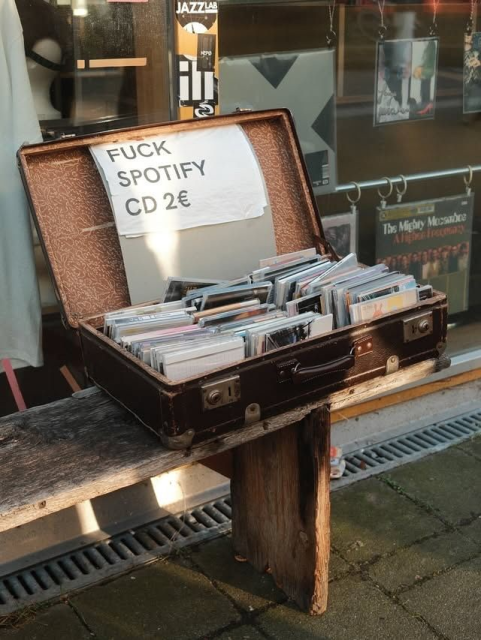 A weathered brown suitcase sitting on a wooden bench outside a shop, filled with numerous CDs in plastic jewel cases. A handwritten sign taped to the inside of the lid reads, "FUCK SPOTIFY CD 2€".