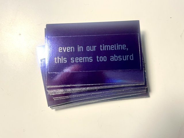 A pile of shiny dark purple stickers with the text “even in our timeline, this seems too absurd”