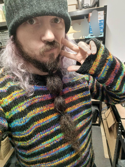 A white masc person with long faded dyed purple/grey hair and a long beard tied with bobbles wearing a black knitted beanie and knitted black jumper with glitchy colourful stripes