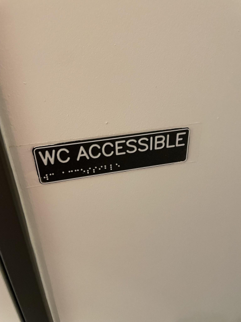Ein 3D-gedrucktes Schild mit der Aufschrift "WC Accessible" in Blockbuchstaben und Braille klebt an einer Wand. Es ist dunkel, die Schrift hell und ein wenig an den Rändern mit Wandfarbe bemalt.