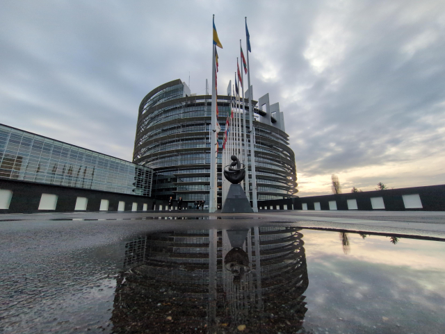 Wide angle picture taken outside the EU Parliament's main entrance, low to the ground.
The building can be seen in its entirety.
In front of it are two parallel lines of flagpoles and a statue.
The weather is quite cloudy, with some light peering through, the ground is still wet from the rain.
A puddle in the foreground shows off a nice reflection of the building and the sky above.