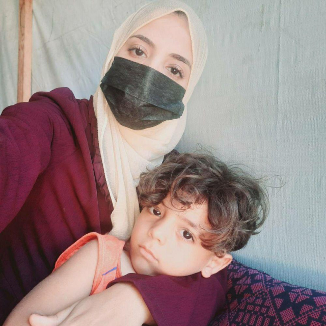A young, 28-year-old woman with fair skin, wearing a medical mask and a headscarf, is holding her four-and-a-half-year-old son, Ibrahim, as they look directly at the camera.