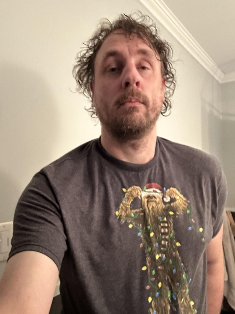 Me wearing a gray T-shirt that has a picture of Chewbacca wearing a red and white Santa hat. He tries to untangle from Christmas lights.