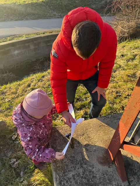 A man and a little girl in winter clothes opening a geocache logbook. It's outside and sunny.