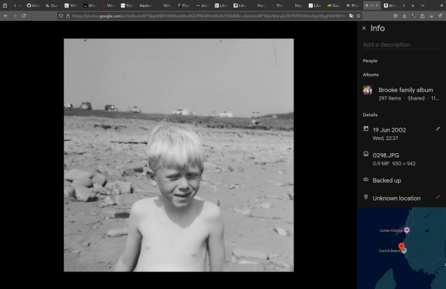 A black and white picture of a small, anxious boy with blond hair, sitting on a stony beach. At the top of the beach, on the grass, a line of cars is visible. The picture is on a Google Photos page, and at the bottom right of the page is a map which shows the exact location where the photo was taken -- and it's right.