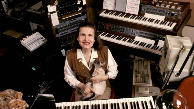 Photo of Wendy Carlos surrounded by keyboards, electronic music equipment, and holding a Sphynx cat in her lap. Wendy is a white woman with shoulder length brown hair with an endearing and awkward smile.