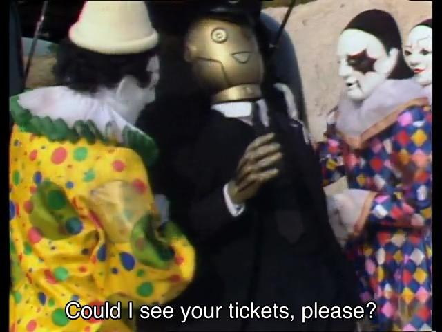 Sylvester McCoy episode. A bunch of clowns are standing around on Segonax, an alien planet that just so happens to look like a dusty quarry. They’re unloading a robot from the back of a van that looks an awful lot like the Kerblam! man, except a bit more ’80s: Lego minifigure-shaped head, more British-looking suit with a tiny tie. The robot has one arm extended out, with a hand that’s shaped far too boney for comfort. It says, “Could I see your tickets, please?”