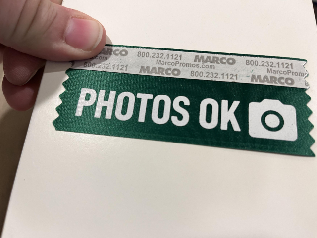 A physical badge ribbon showing “photos OK” as in the proof
