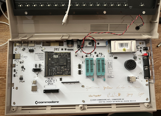 The Commodore 64 Ultimate with the top of the case removed. The main board is all white with golden signatures of the creators on it. The brains of the board is a single highly integrated daughter board attached to the main board. There are two large empty slots available to use genuine legacy SID chips from the 80s. There's a small speaker. There are various connectors and some other modern components sprinkled around, but largely it's pretty empty.