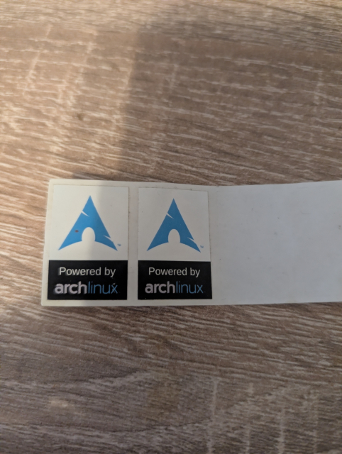 Dvě samolepky s logem Arch Linuxu a nápisem „Powered by archlinux“ leží vedle sebe na dřevěném stole.
