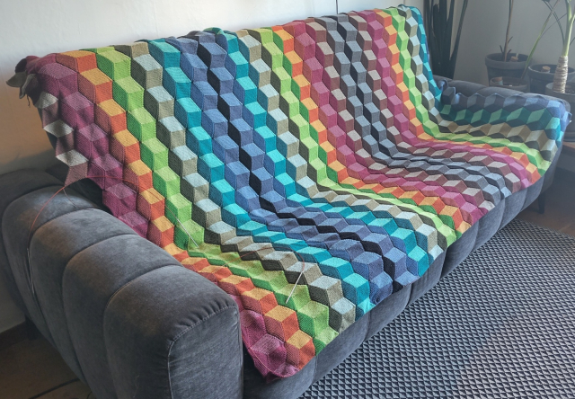 Photo of an almost done knitted blanket in 23 different colours. The pattern is a bit hard to look at. It can be seen as depictions of cubes, or just rows of interlocking parallelograms.