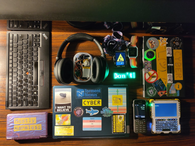 Photo showing a number of personal electronic devices including a Bluetooth Thinkpad keyboard, a couple of laptops adorned with Chaos-adjacent stickers, a Tanmatsu, a DECT phone (no base station, awww), a Mysterious Clamshell Device, some headphones, a BangleJS watch, a Pimoroni Tufty running a CO2 monitoring app, a FOSSAsia name badge showing the word "Don't" and a little tub full of the smaller personal electronic devices....