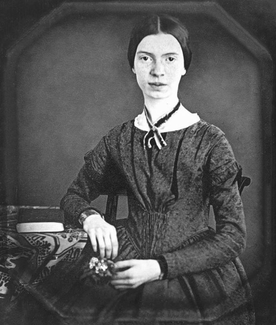 Emily Dickinson seated by a table with a tablecloth and two books stacked on top.