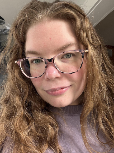 A photo of Ashe wearing a purple sweatshirt. Her blonde hair is down and some of it is wavy/curly and some of it is straight. She has a light smile and looks into the camera.