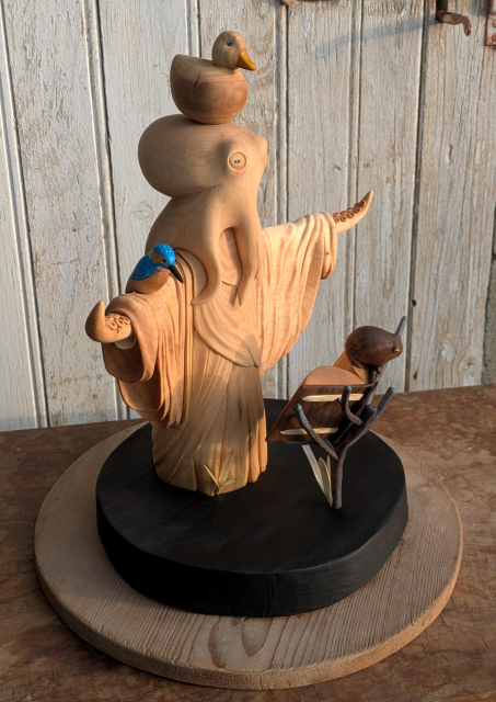 Woodcarved humanoid octopus wearing a robe, arms wide open, in front of a book standing on branches. On the book, a frog. On the right arm, a kingfisher, and on the top of the head of the octopus, a round duck. The base is burnt, on that angle it is not really obvious but is water with a slight wave form arround the octopus. On the background, an old door with old white paint.