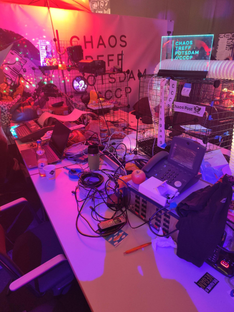 Chaotischer Tisch vom Chaostreff Potsdam. Auf dem Tisch viel Technik, u.a. ein ISDN-Verteiler und ein ISDN-Telefon, eine Erika-Schreibmaschine, mehrere Laptops. Im Hintergrund ist unser Banner zu sehen