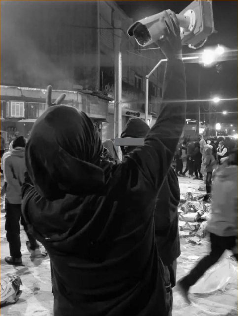 A picture of a person in Hijab who holds a surveillance camera in her right hand letting the camera point at them.  But the camera is disconnected and fully in the person hands, blinding whoever might try to surveill.  The person is in the near a street crossing and there are people everywhere on the street, in a protest/uprising it appears.  The picture was, as far as we could verify, taken in Abadnan on January 7th.  See https://en.wikipedia.org/wiki/Abdanan