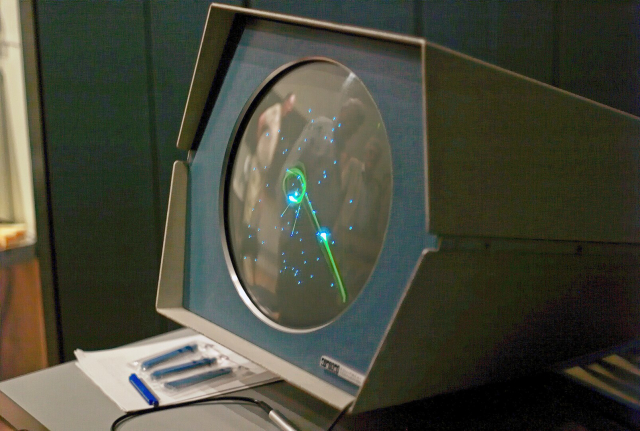 A close-up of a vintage PDP-1 computer display running *Spacewar!*, shown on a circular CRT screen inside an angular metal housing. Green vector lines and glowing dots form a starfield with two small spacecraft maneuvering around a central gravity well. The room is dim, with the machine sitting on a table alongside papers and pens, emphasizing the early, experimental era of computer gaming.