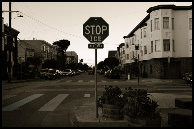 A monochrome picture taken late yesterday afternoon, at an intersection in the Mission District, San Francisco. It shows a stop sign rising up from a corner of the intersection. The sign is set against the sky, with 100-year-old buildings on either side. Traffic and park cars line the street. 

This is 2026, so the sign has been enhanced with spray painted block letters. It now reads: 

STOP
ICE