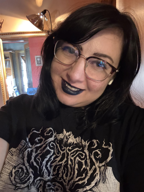 A selfie of me smiling at the camera. I have jet black hair that is shoulder length and side swept, and I’m wearing black lipstick and a black t-shirt that has the logo for the band Antrisch