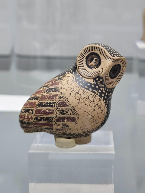 A vessel in the shape of an owl, displayed on a transparent pedestal. The figurine is intricately painted in black, red, and beige, with finely detailed feathers, large round eyes, and a slightly tilted head.