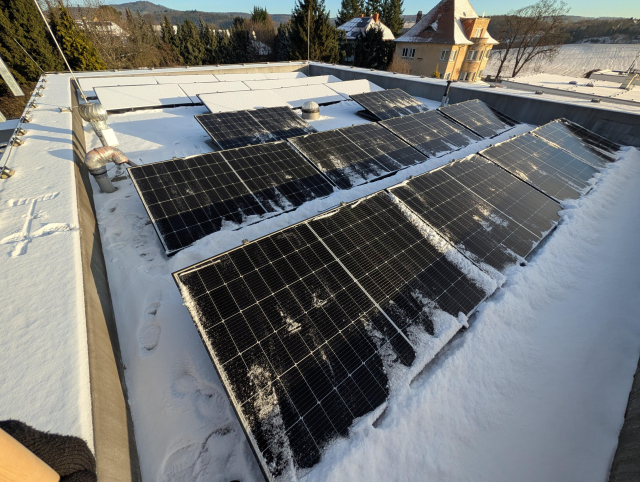 A flat roof with 21 solar panels. Half of them are clean, half of them are covered in snow.