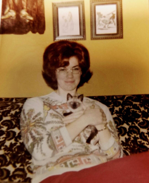 Mom, aged 30, at home, happily cradling a Siamese kitten. She is wearing an abstract print top with coral pants. The couch has that heavy raised felt design on the upholstery, dark moss green on a green background. A couple pictures and a hammered copper hanging are on the wall behind her. She sports a flip hairdo and the new acetate frame glasses that had come out.