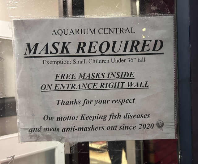 A printed paper sign taped to the front door of the shop. Black text reads:

AQUARIUM CENTRAL

MASK REQUIRED

Exemption: Small Children Under 36" tall

FREE MASKS INSIDE
ON ENTRANCE RIGHT WALL
Thanks for your respect

Our motto: Keeping fish diseases and mean anti-maskers out since 2020
