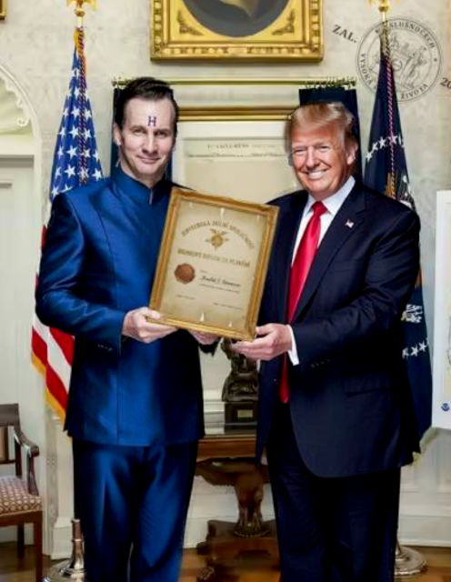 A picture from the Oval Office where a man with "H" on his forehead and in a blue suit, a character from the British sitcom Red Dwarf, is giving a framed certificate to Donald J. Trump. They're both smiling.