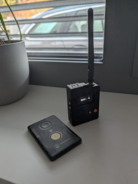 Two Meshcode devices in front of a window. One has a form factor of a credit card, it's called SenseCAP T1000-E, the other is black and has a form factor of a walkie talkie, its name is Wio Tracker L1 Pro.