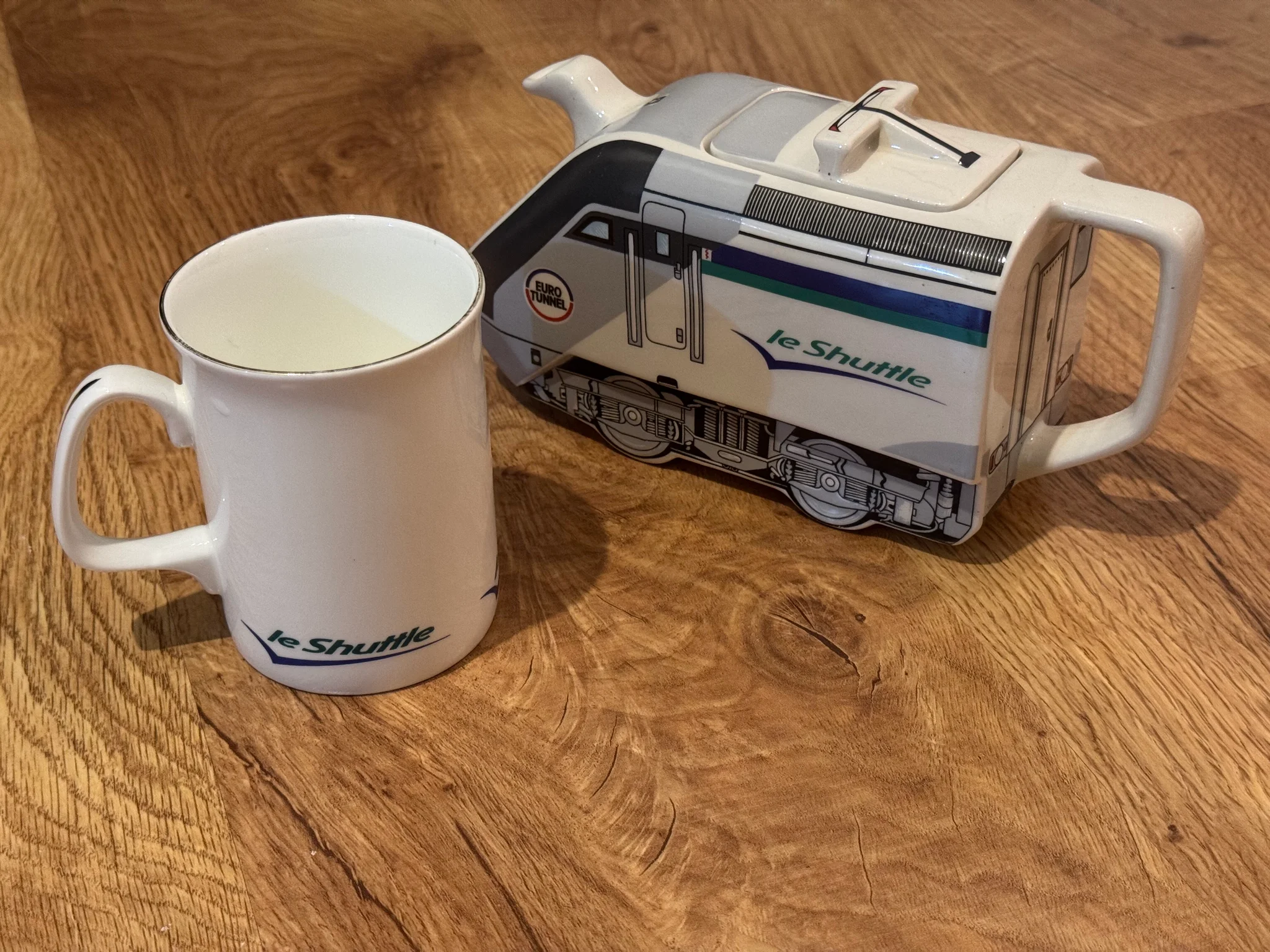 A teapot shaped like a Eurotunnel Class 9 electric locomotive, complete with the lid featuring a pantograph, and the spout coming out of the front of the locomotive. It is next to a Le Shuttle mug.
