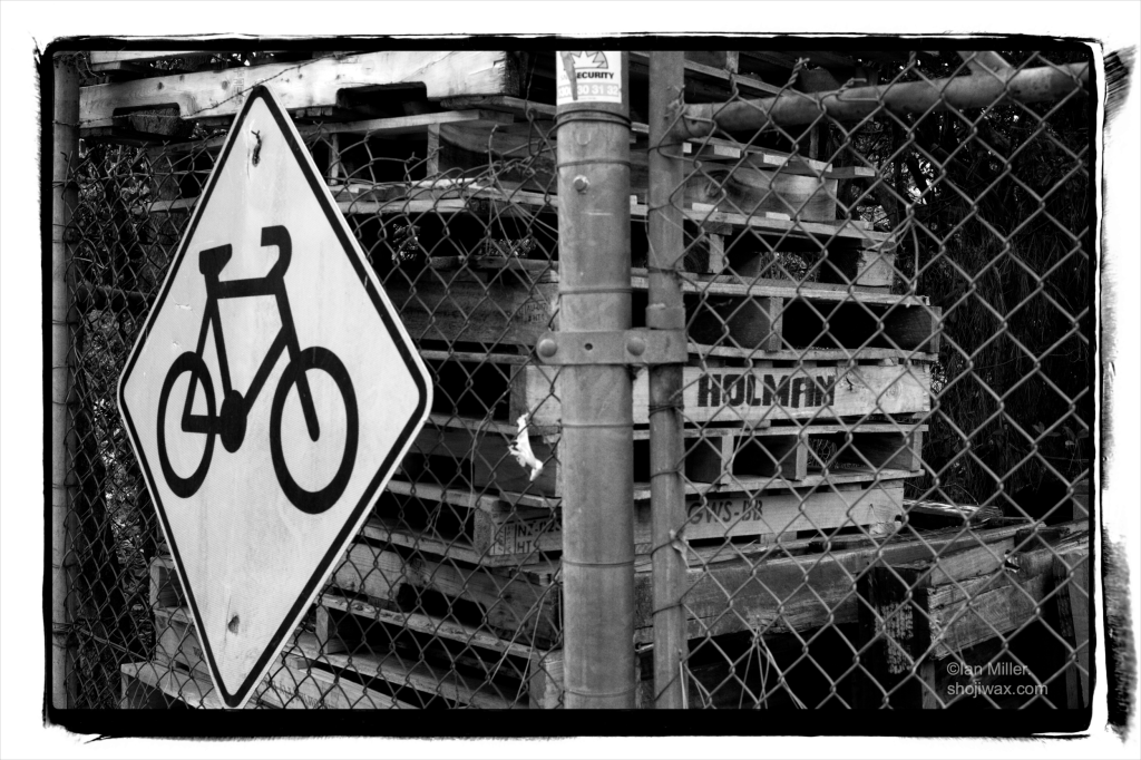 Monochrome photo of a sign with a bike symbol on it mounted to a wire fence. Behind the fence can be seen a stack of wooden palates.