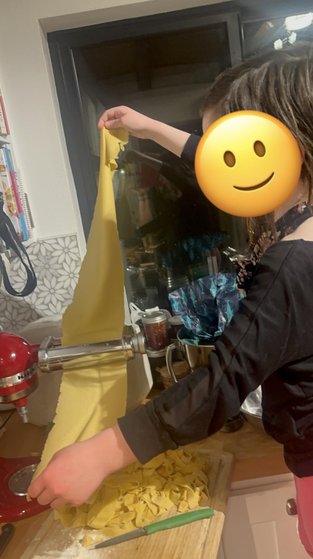 A photo of a child standing on a chair feeding a sheet of pasta through a pasta maker attached to a kitchen-aid mixer. The kid’s face is obscured by a smiling emoji but take my word for it that she’s adorable. A pile of already-cut pappardelle are on a cutting board below the mixer. The scene looks warm and happy.
