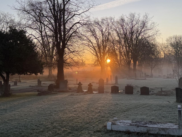 Picture shows a cold frosty morning in a cemetery. The grass has a white frost layer leading to the silhouettes of several tombstones. The outline of tall leafless trees are lit from behind as a low orange sun begins to rise. The glow from the suns rays illuminates the frost giving the illusion of fog rising from the ground. It gives a spooky feel.