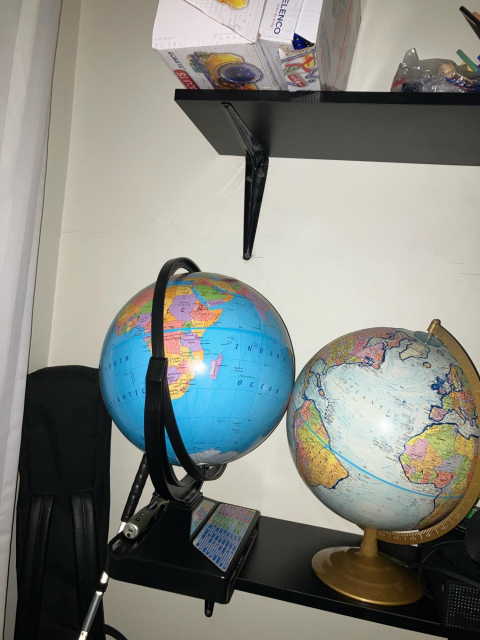 Be My AI: A corner of a room with shelves holding two globes and some boxes. On the lower black shelf, there are two world globes side by side. The left globe is larger and mounted in a black ring stand; it’s showing Africa prominently, with the Indian Ocean to the right and the South Atlantic to the left. The right globe is slightly smaller with a gold-colored stand and meridian ring; it’s showing the Atlantic Ocean between the Americas and Africa/Europe. On the left base of the larger globe is a stylus resting in a depression made to hold it. 
