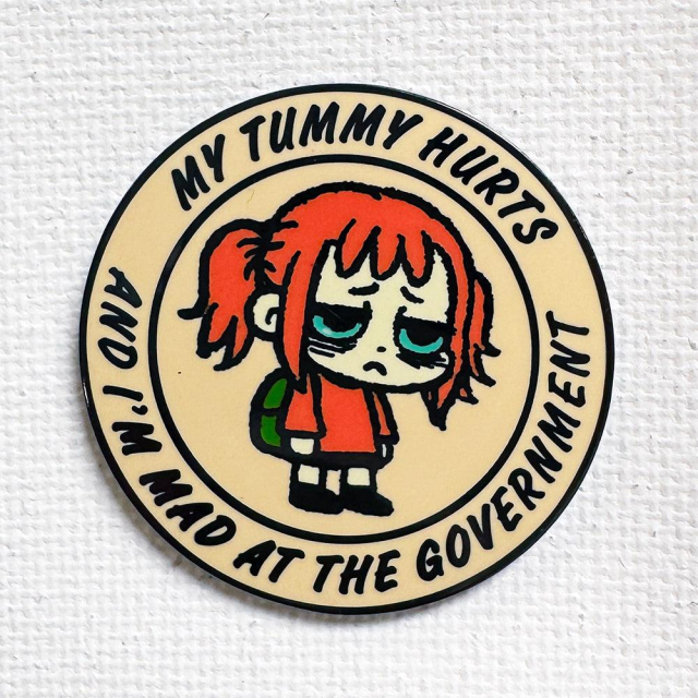A circular enamel pin featuring a cartoon girl with messy red hair and tired, dark-circled eyes. She wears an orange shirt and a green backpack. Around the border, bold black text reads: "MY TUMMY HURTS AND I'M MAD AT THE GOVERNMENT."