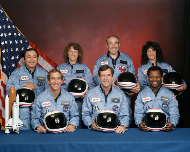 An official NASA photo of the seven astronauts of the STS-51L Challenger space shuttle mission. They are all smiling, in light blue flight suits with crash helmet style protective headgear, seen against a reddish backdrop with a US flag. Three are seated with the other four standing behind.
