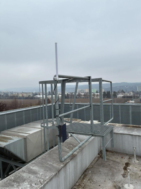 An antenna and a black device with a solar panel attached to a metal construction the the top of a building.