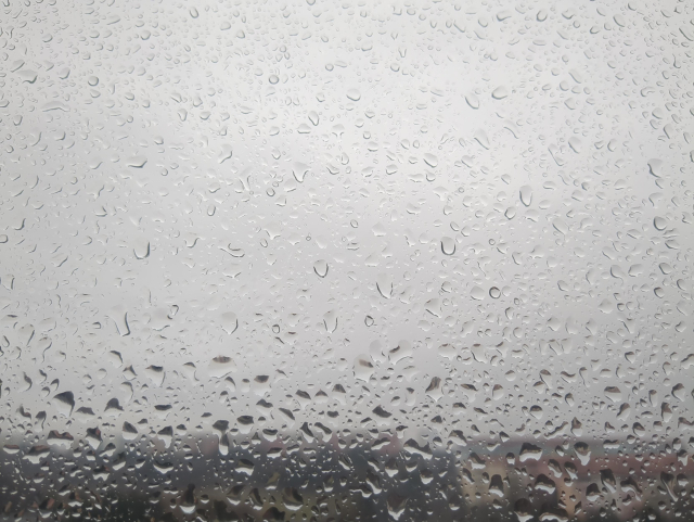 Fotka okenní tabule pokryté kapkami, ve spodní pětině je vidět nezaostřený horizont, zbytek pozadí je šedobílý. 